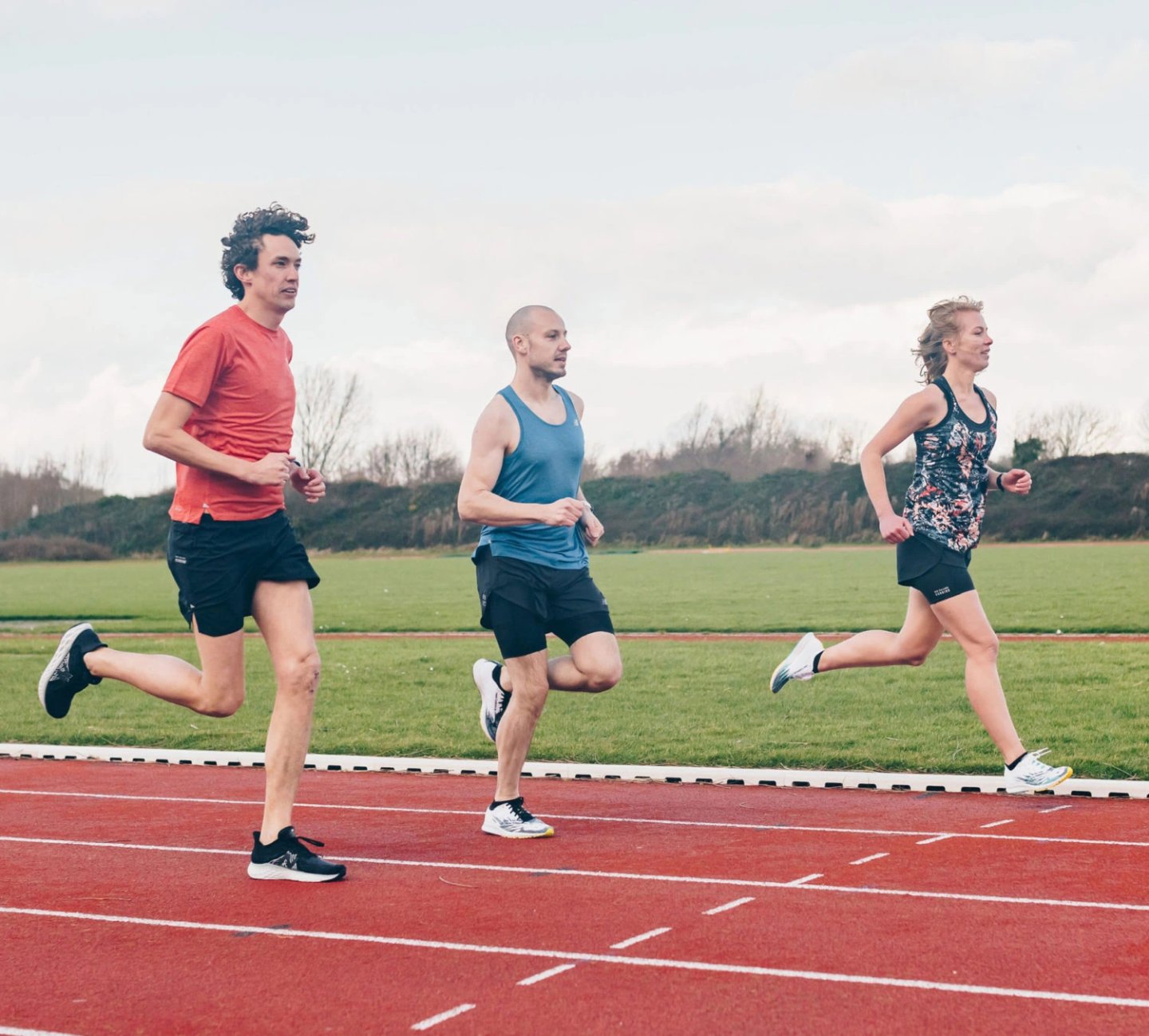 Two male runners and one female runner training on a red athletics track, wearing black shorts and performance running tops.