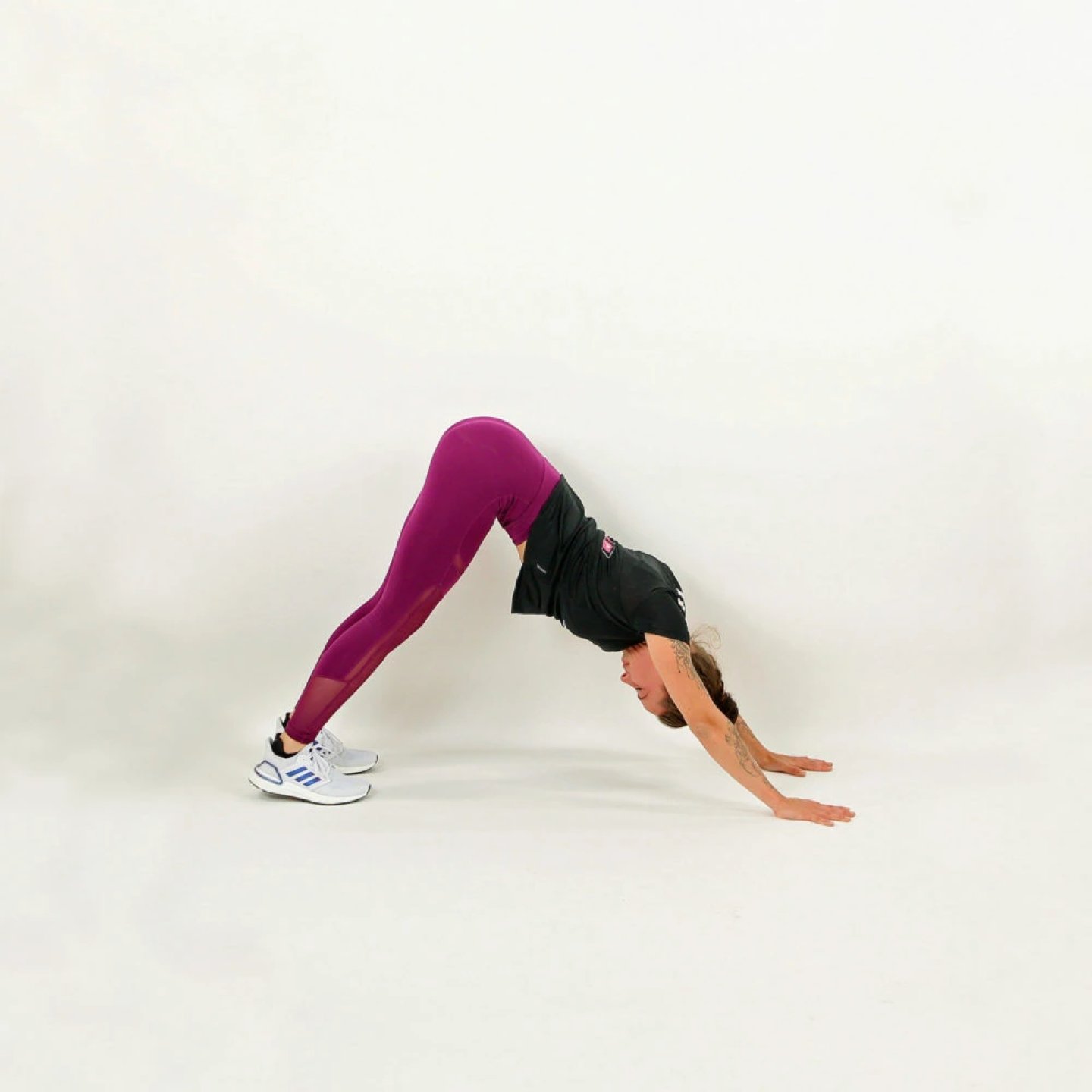 Woman in purple running tights and black top performing a downward dog stretch on a white studio background.
