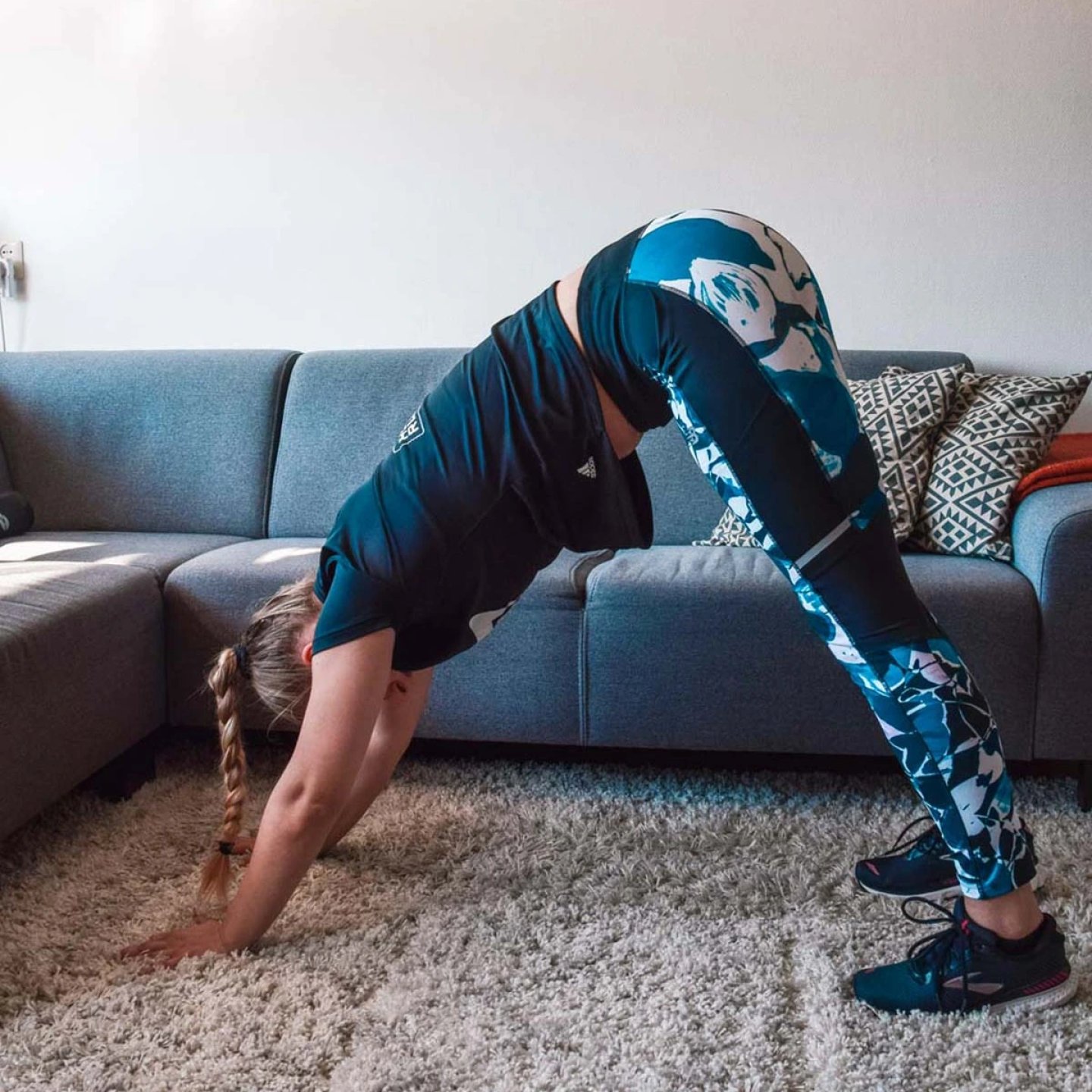 Woman in blue patterned leggings and dark Adidas top performing a downward dog stretch on a carpet at home.