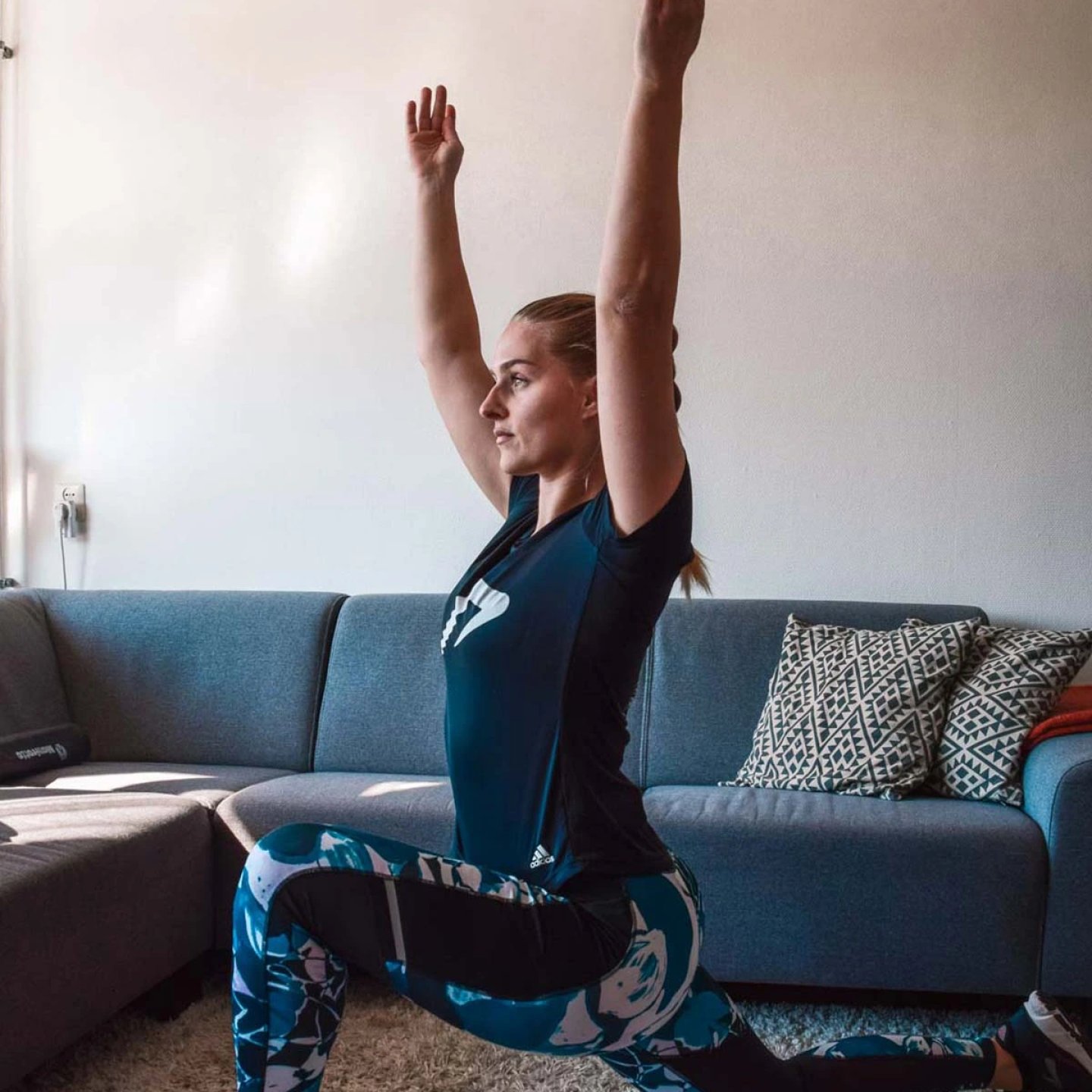 Woman in blue Adidas tights and navy t-shirt performing a deep kneeling lunge stretch with arms raised overhead indoors.