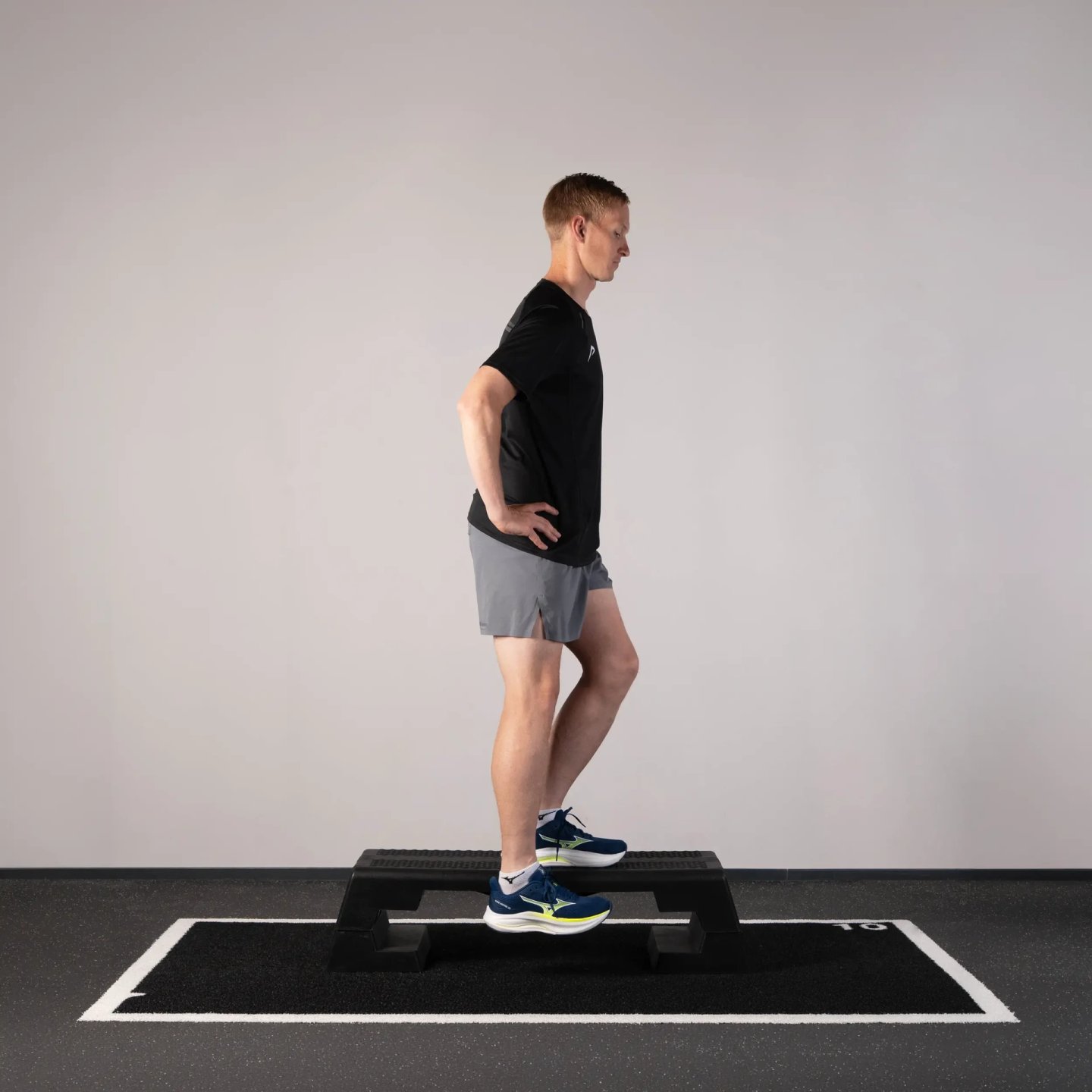 Male athlete in black tee and grey shorts performing a step-down exercise on a gym step platform wearing blue and yellow running shoes.