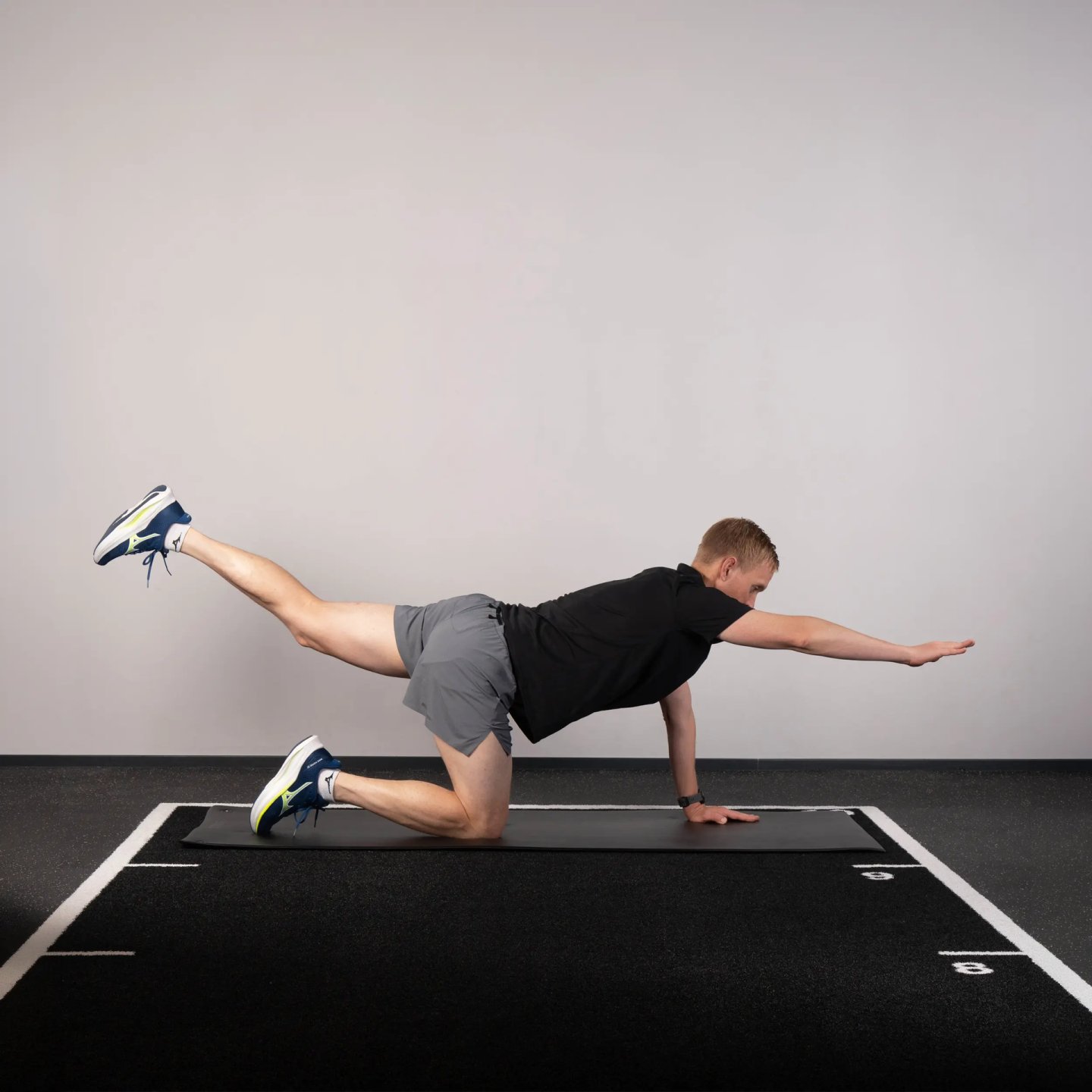 Male runner performing a Superman exercise on a black gym mat, extending opposite arm and leg while wearing Mizuno trainers.