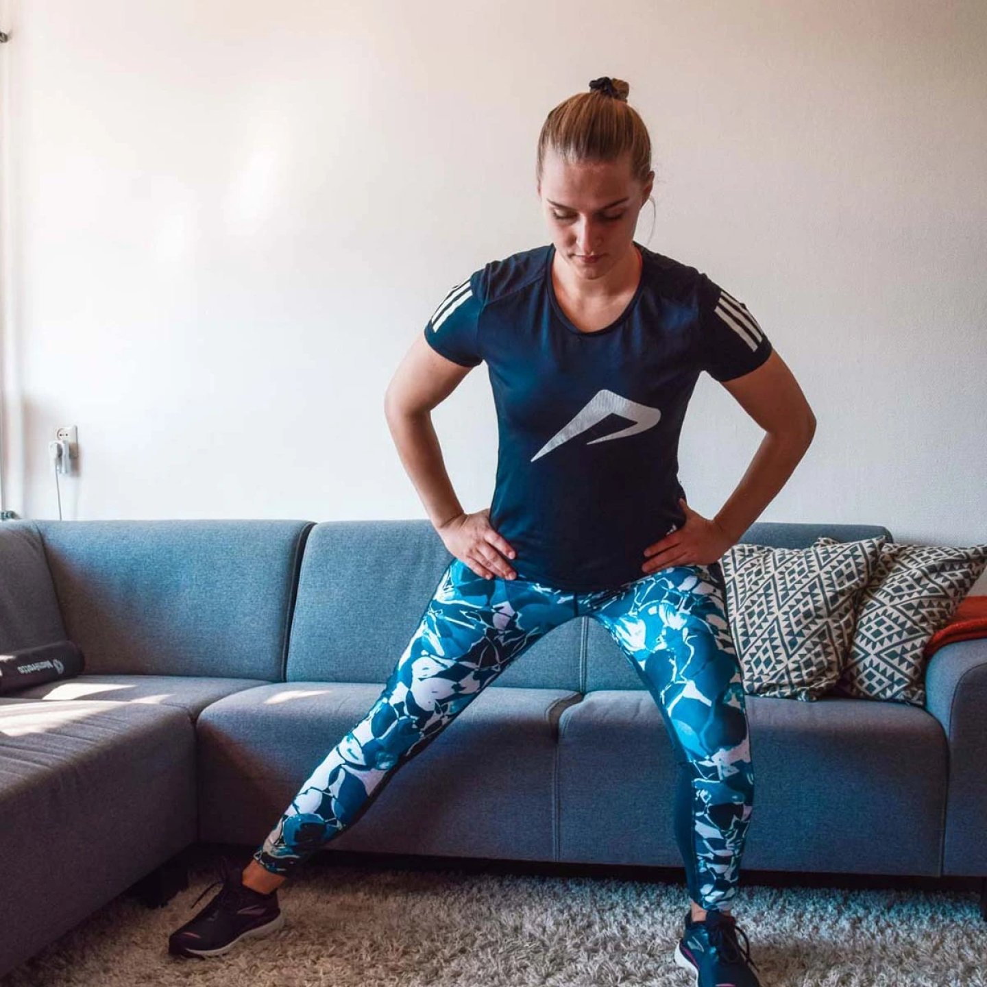 Woman in blue floral-print running tights and navy Adidas training tee performing a side lunge in a living room.