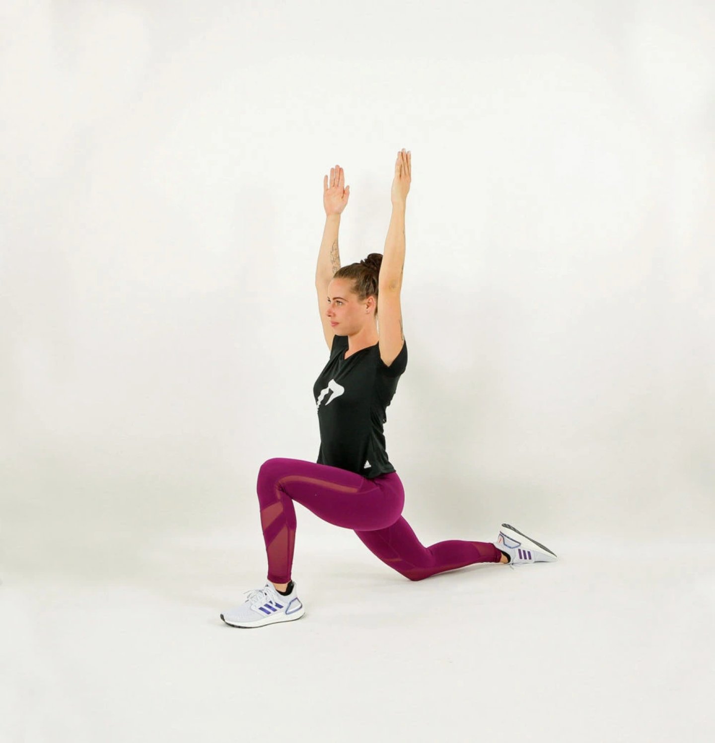 Woman in purple leggings and black tee performing a kneeling lunge with arms raised overhead, wearing white Adidas trainers.