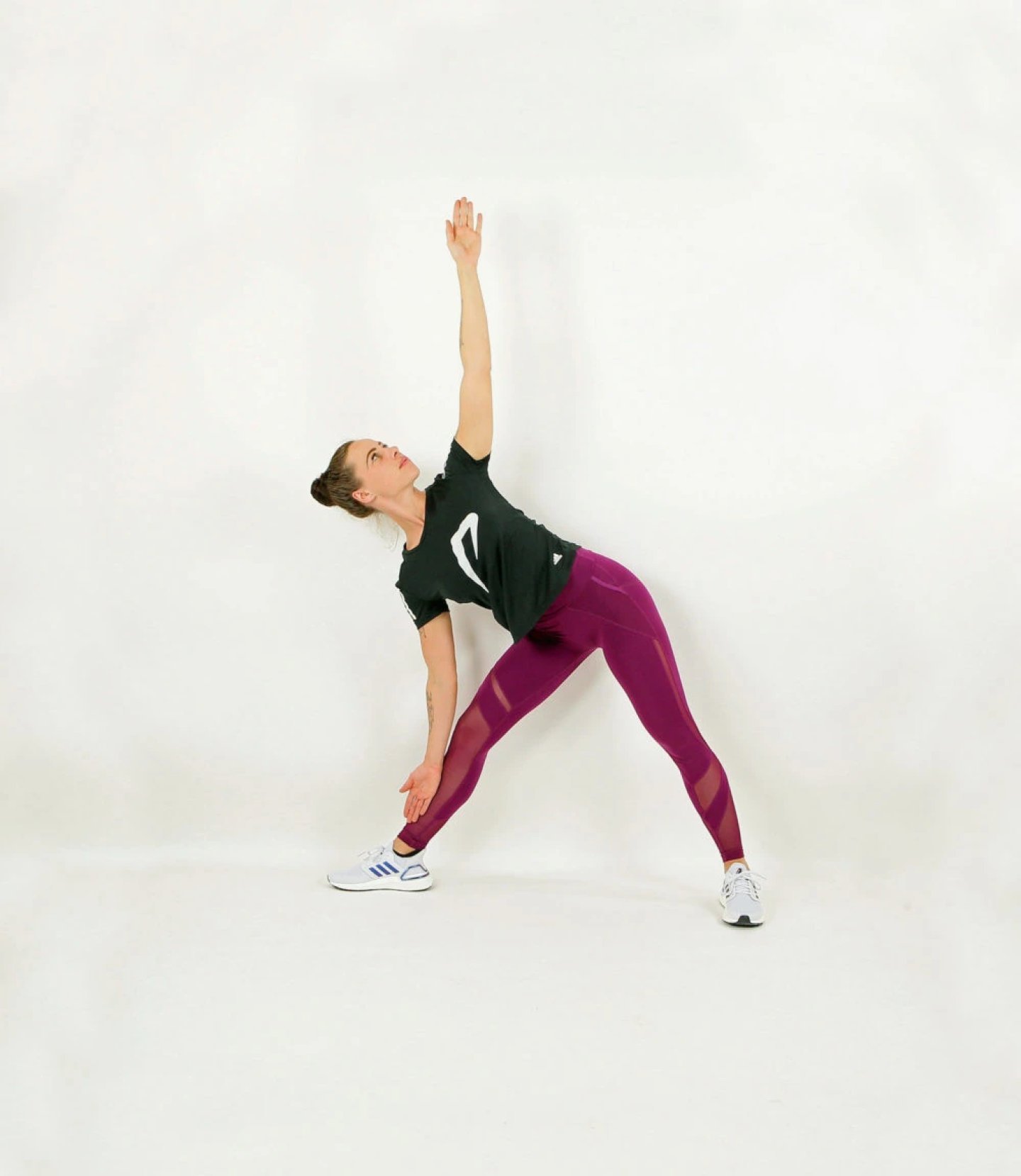 Woman in purple mesh leggings and black adidas tee performing triangle pose stretch on white background