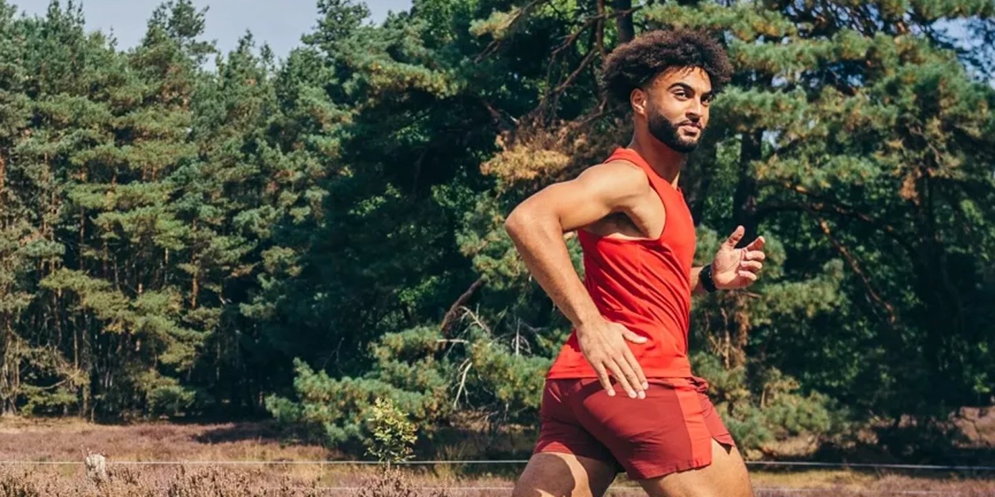 Man wearing Saucony trail running red vest and shorts running outdoors along a forest path in sunny conditions.