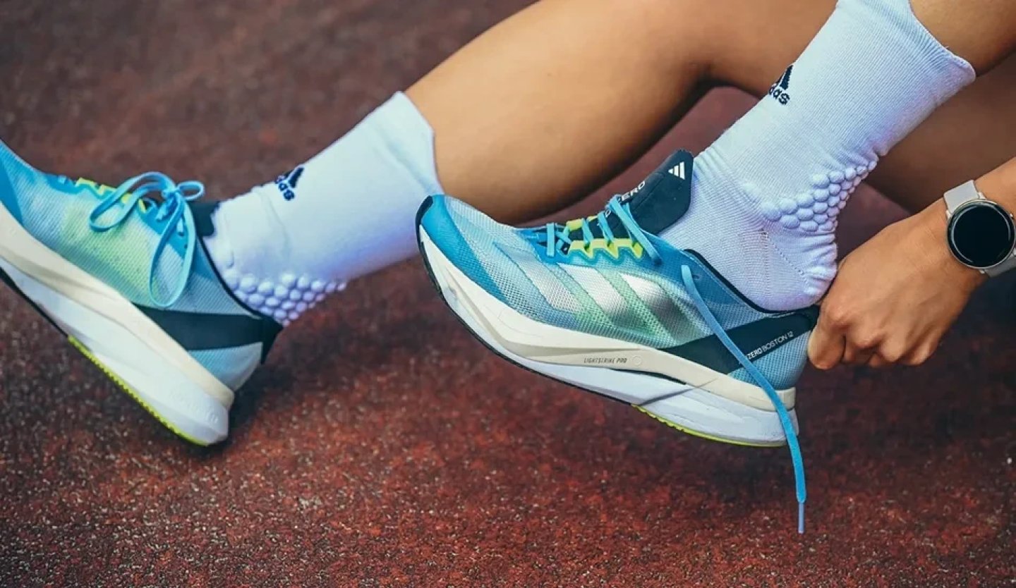 Runner lacing up adidas Adizero Boston running shoes in aqua blue gradient with Lightstrike Pro midsole on a red track