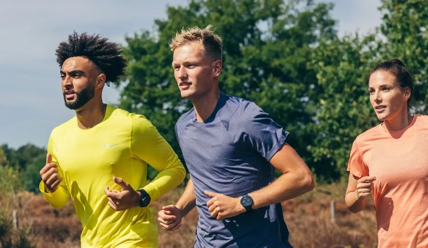 Three runners wearing Saucony trail running apparel jogging outdoors on a sunny day with trees and heathland in the background.