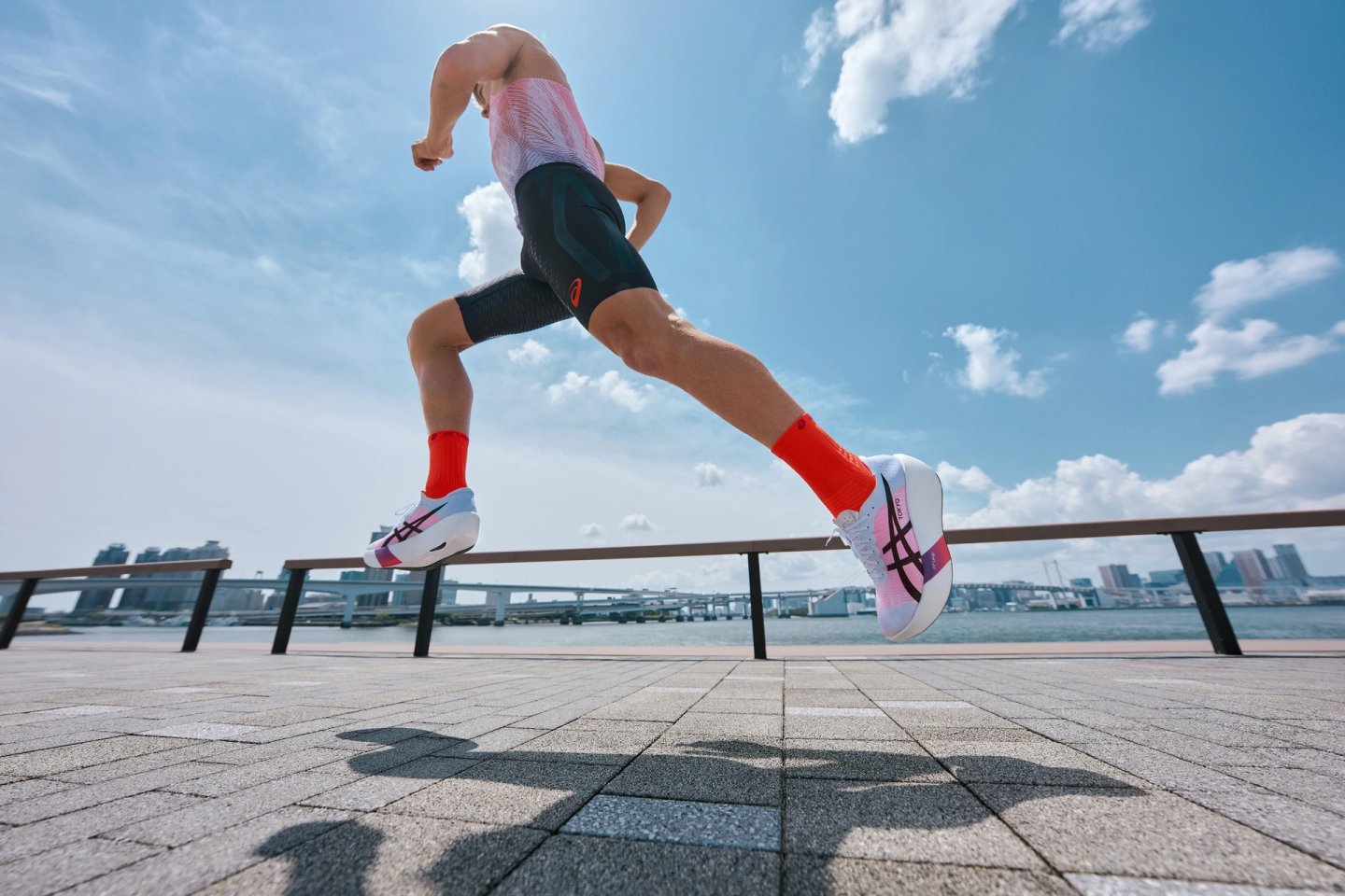 Male runner leaping mid-stride wearing Asics Tokyo 2025 white racing shoes, red socks, and black compression kit by a waterfront.