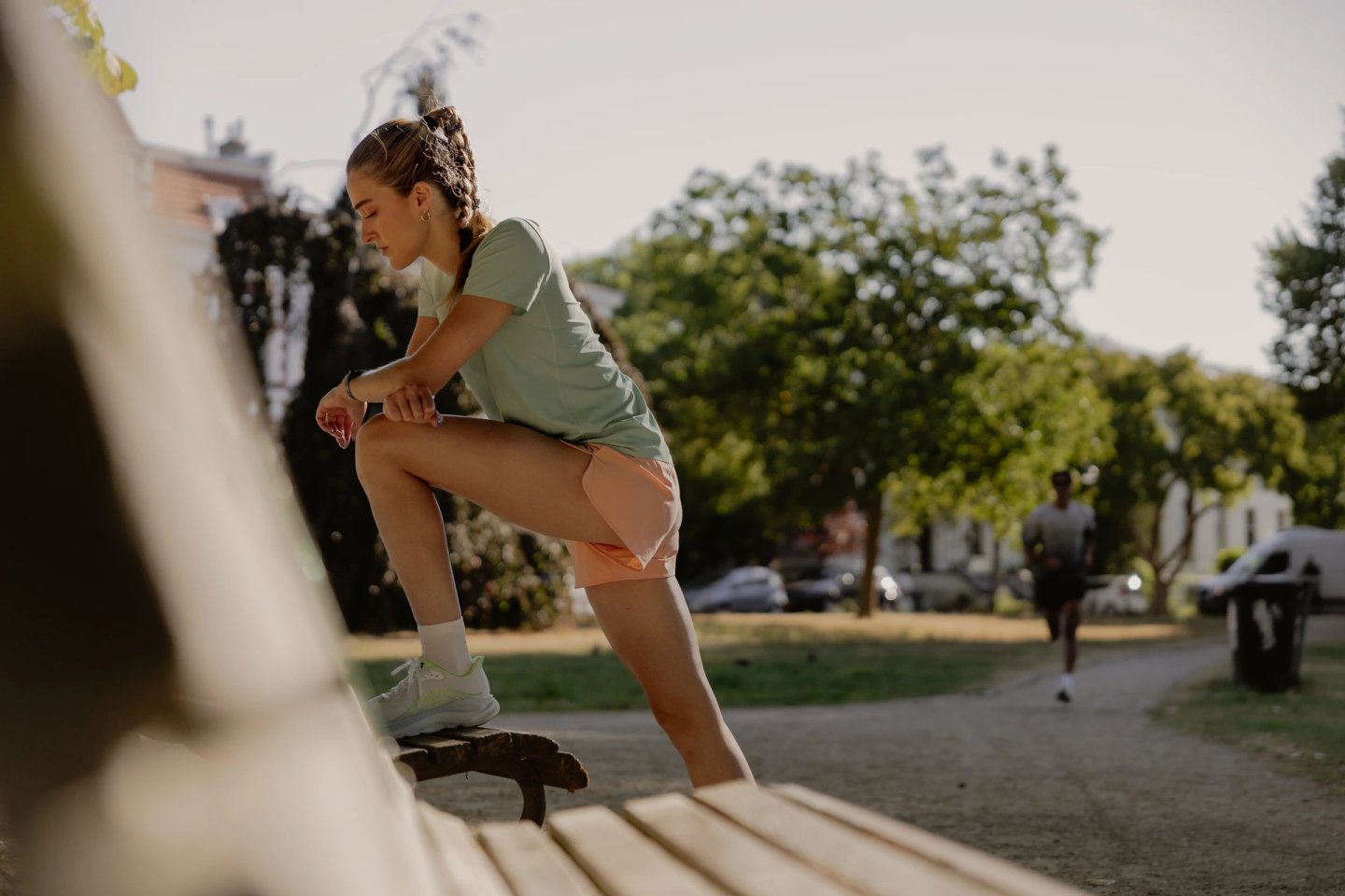 Woman in mint running t-shirt and peach shorts resting foot on park bench, checking smartwatch outdoors