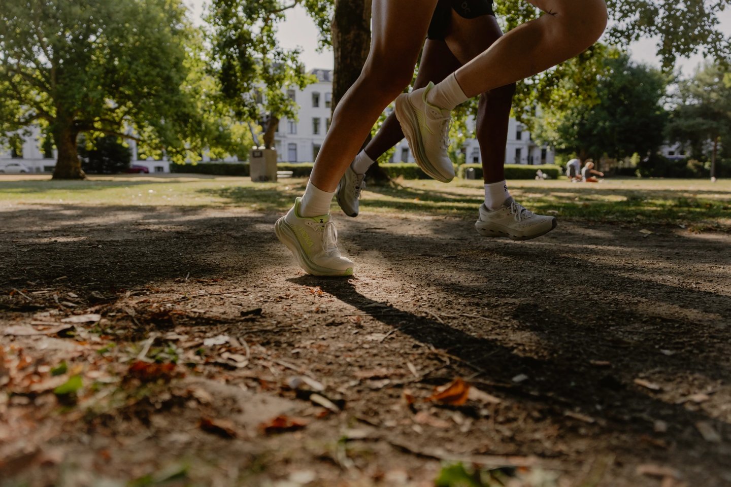 Low-angle shot of two runners wearing white running shoes on a dirt park path in summer sunlight.