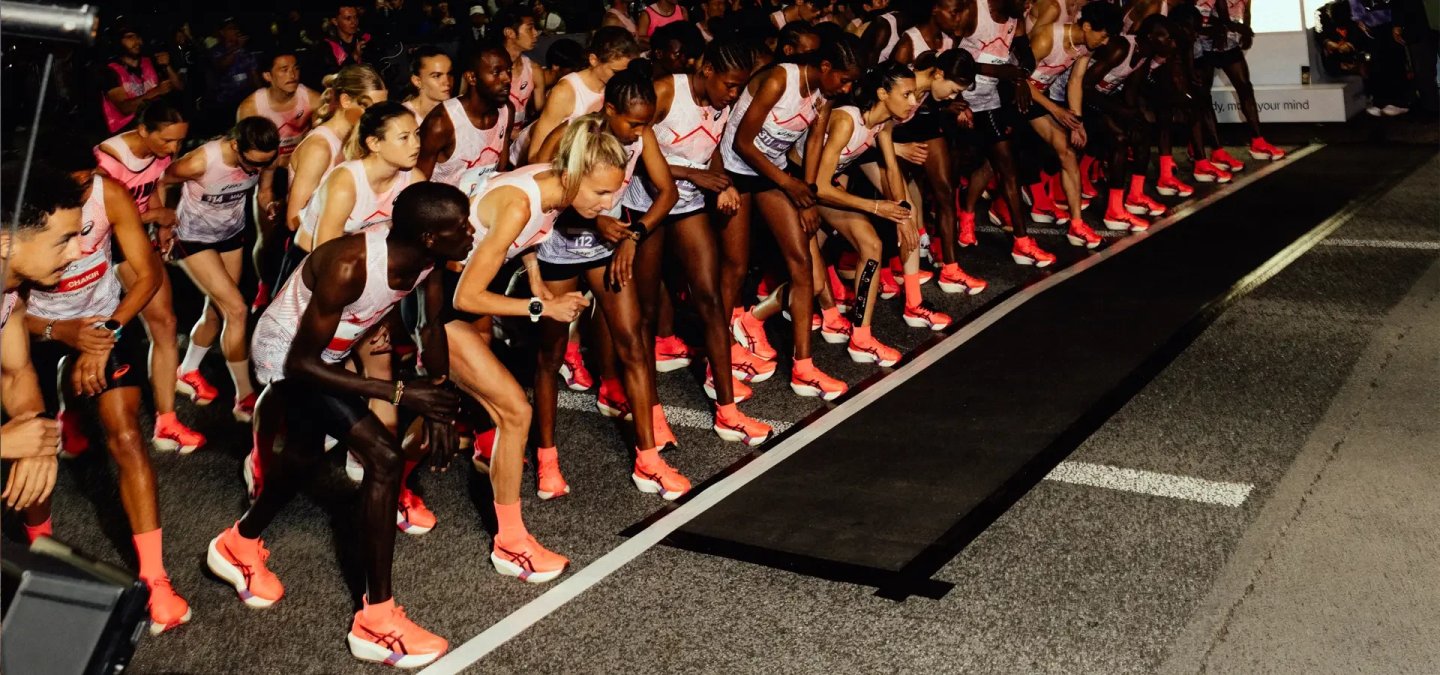 Large group of elite runners in Asics white and pink race vests and coral racing shoes crouched at a start line at night