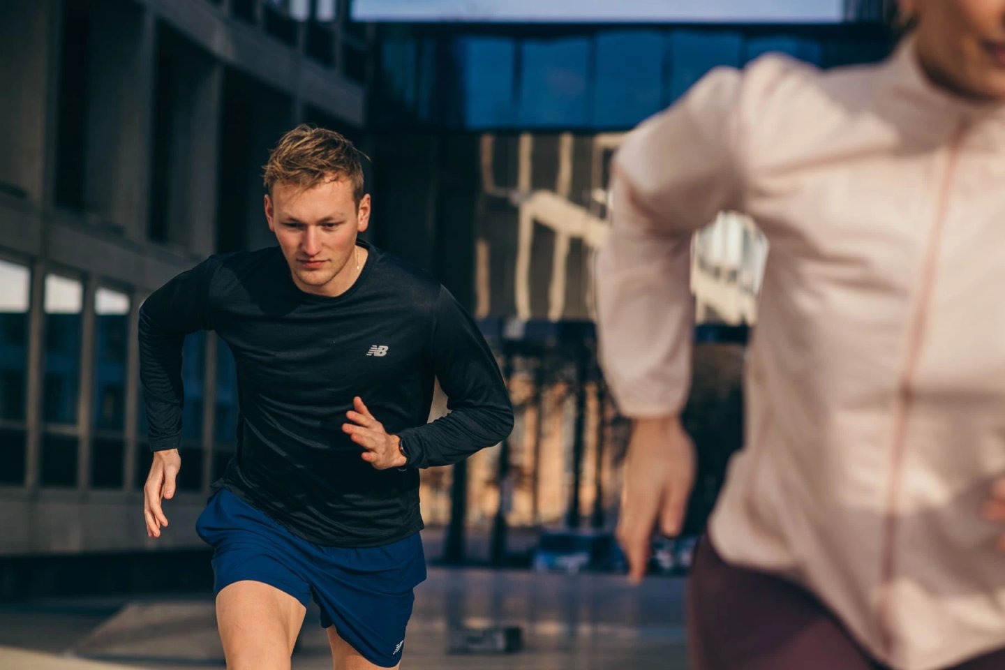 Man running in a black New Balance long sleeve top and blue shorts through an urban street setting