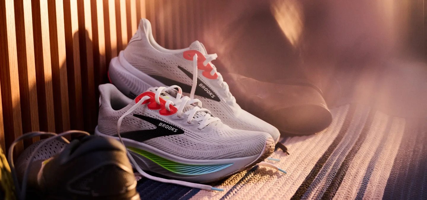 Two Brooks Ghost running shoes in white with red lace accents, green and blue midsole detailing, placed on a textured surface
