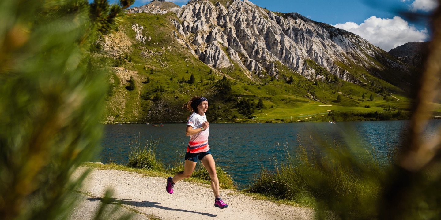 Woman trail running on a gravel path beside a blue alpine lake with rocky mountain peaks in the background