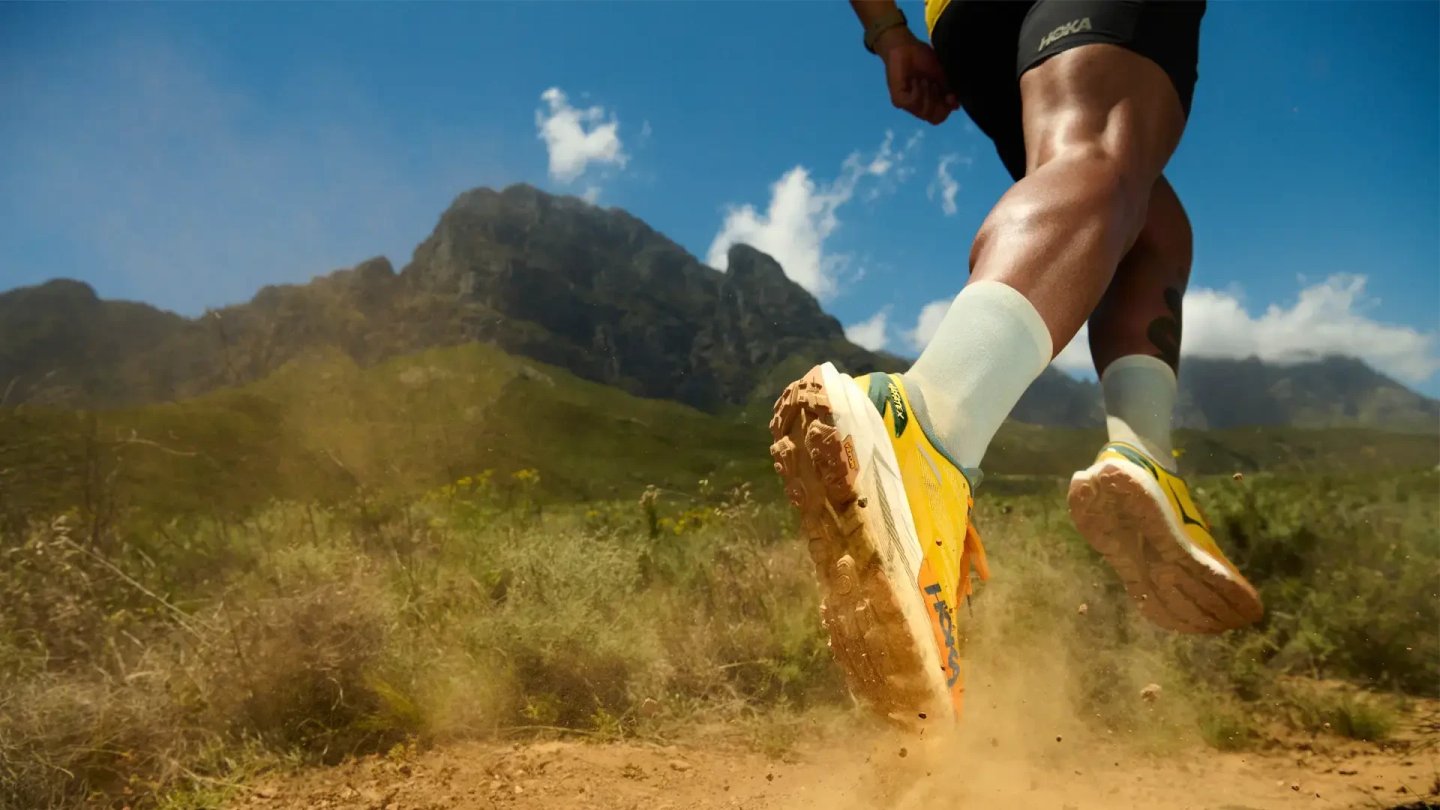 Close-up of runner wearing yellow Hoka trail running shoes kicking up dust on a dry mountain dirt trail
