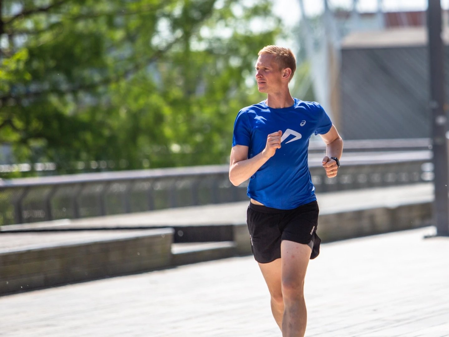 Male runner in blue ASICS t-shirt and black shorts running on a sunny urban boardwalk with trees in background