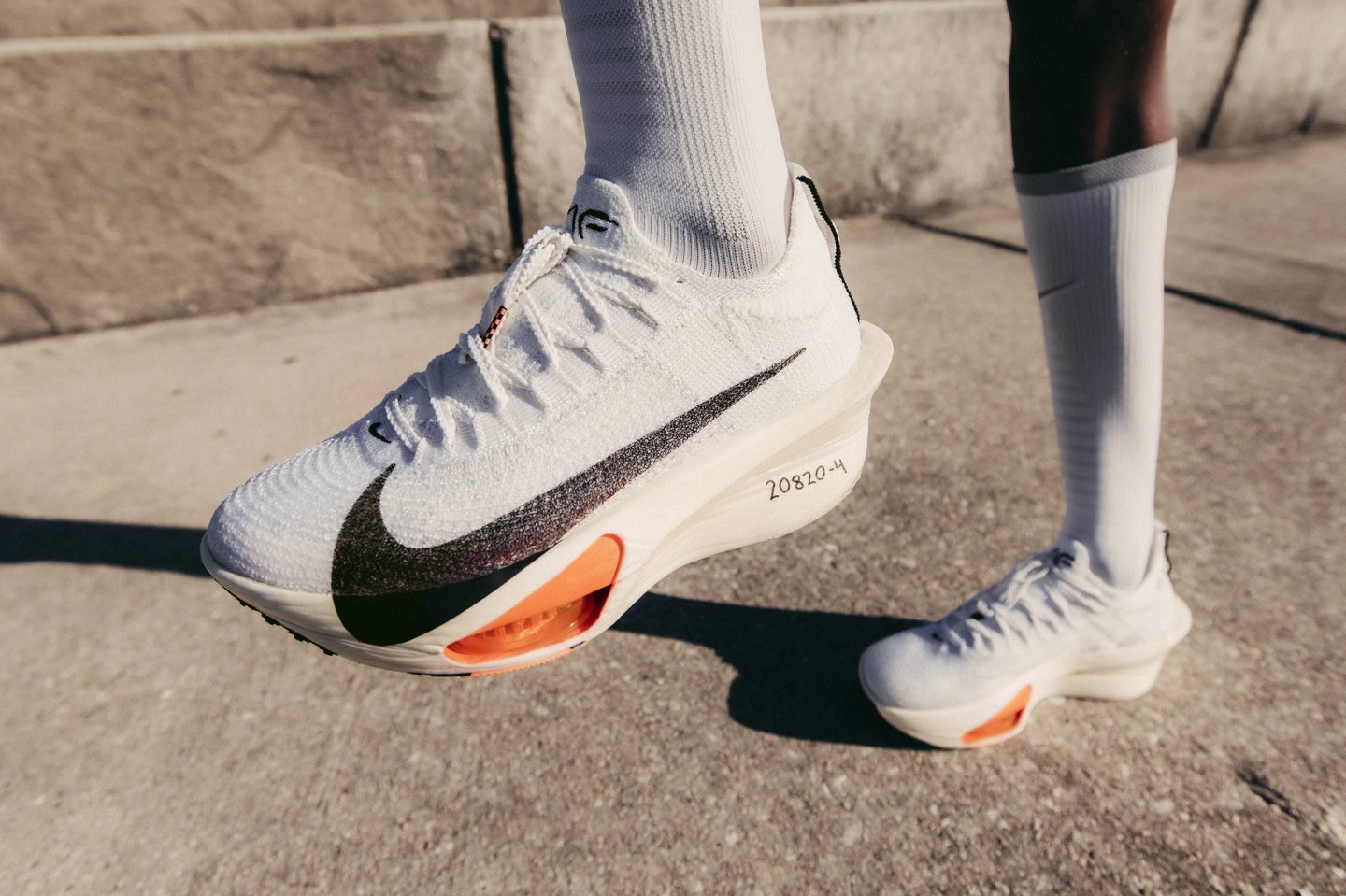 Nike Alphafly 3 Proto running shoe in white with black Swoosh and orange Air Pod cushioning, worn on concrete pavement.