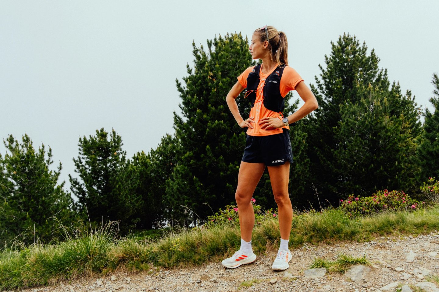 Woman in adidas Terrex orange tee, black skort and hydration vest standing on a gravel mountain trail surrounded by pine trees.