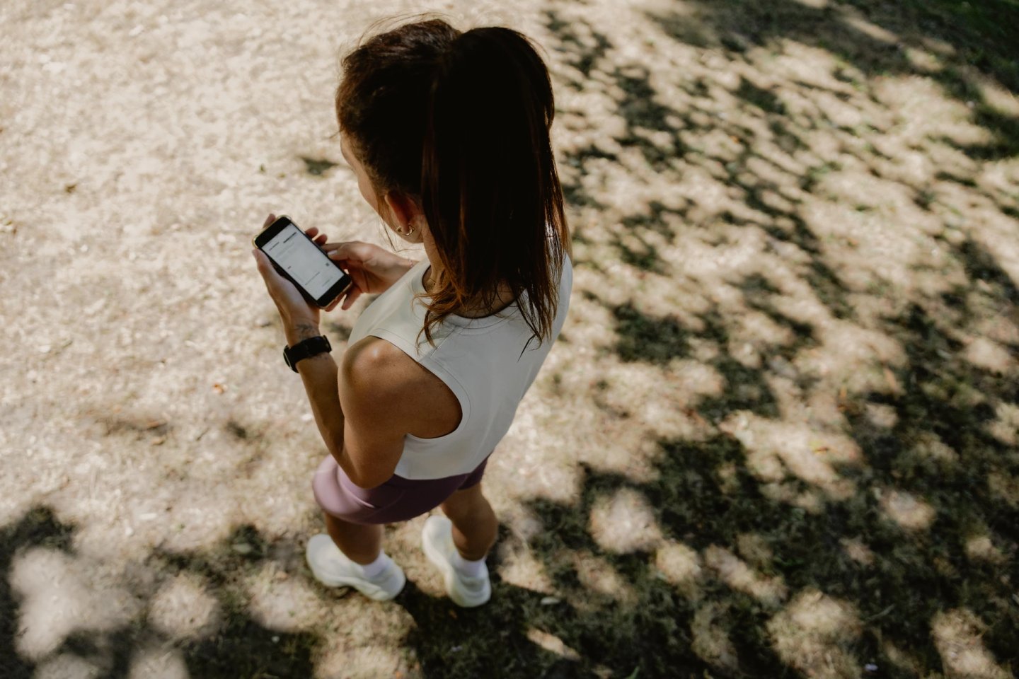 Overhead view of a female runner in white tank top and mauve shorts checking her smartphone in a park