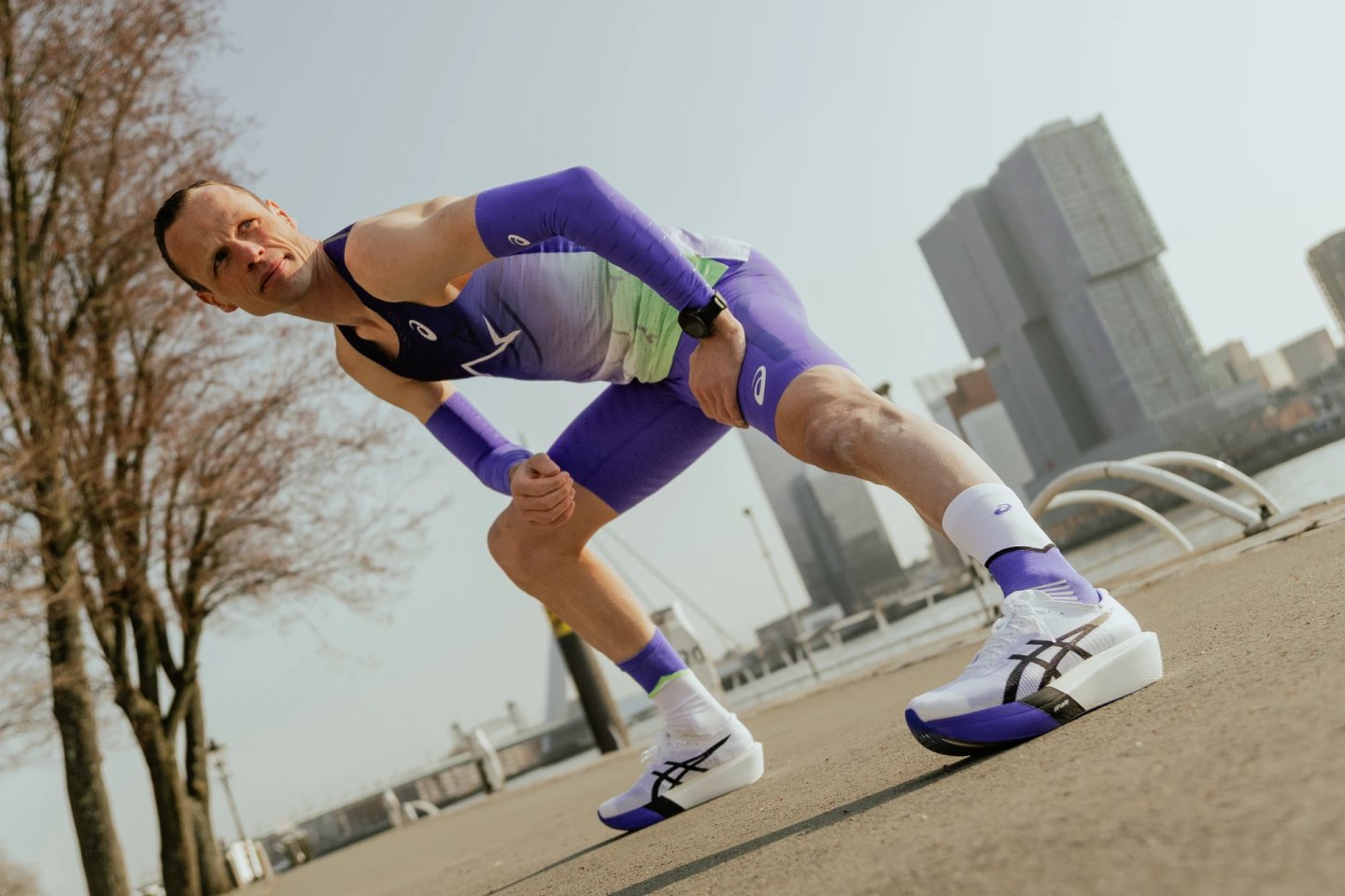 Male runner in purple ASICS race suit and white ASICS carbon-plate racing shoes in aggressive sprint stance by Rotterdam waterfront.