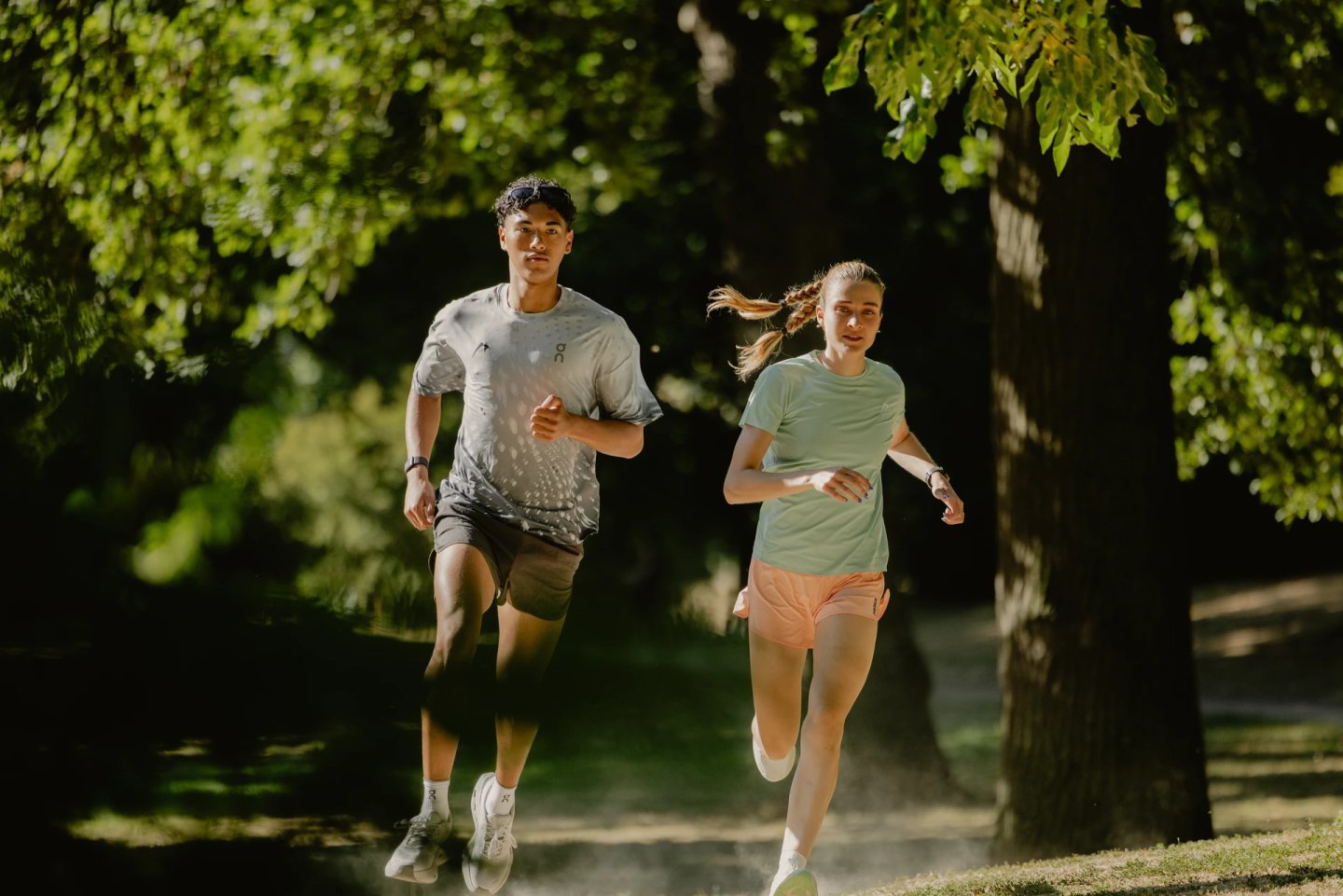 Man in grey On running tee and woman in mint green tee and peach shorts running together on a shaded park path.