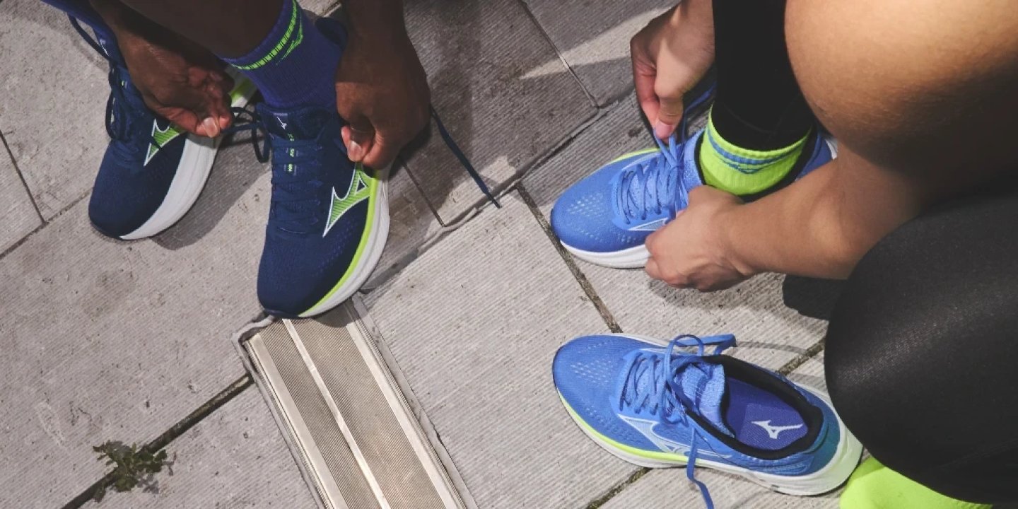 Two runners tying laces on Mizuno Wave Rider shoes in navy green and blue colourways on urban pavement