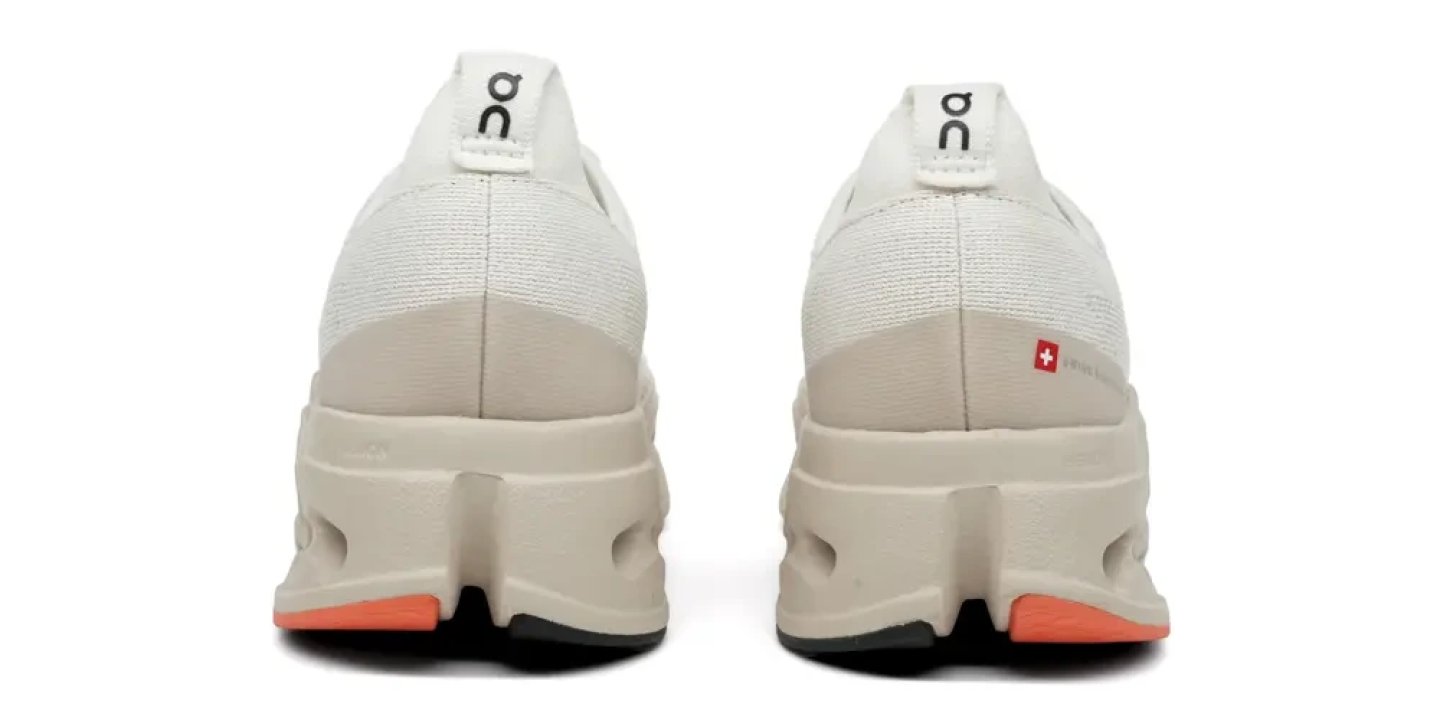 Rear view of On Cloudmonster running shoes in white with beige stacked midsole, coral outsole and Swiss flag detail