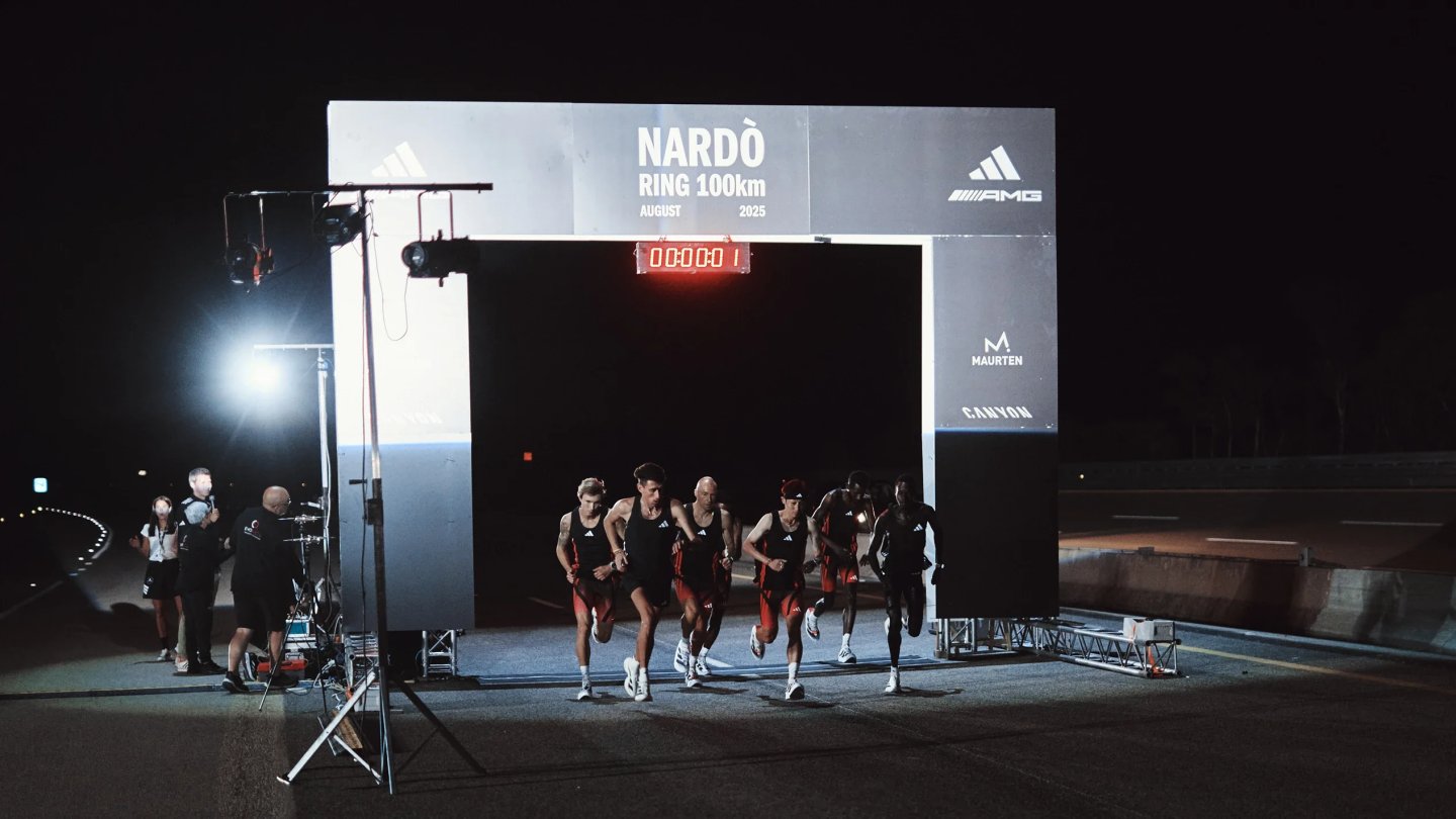 Elite runners in adidas race kits sprint through the illuminated start arch of the Nardò Ring 100km night race, August 2025.