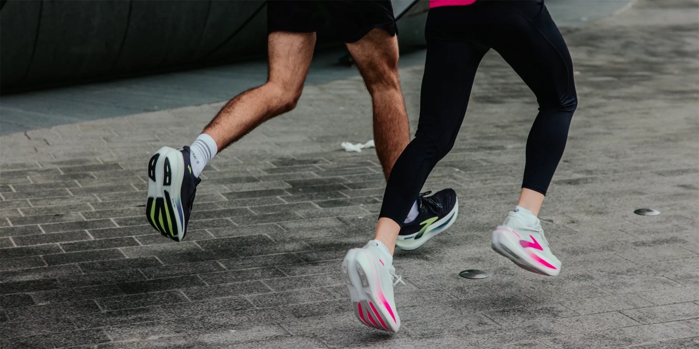 Close-up of two runners' legs mid-stride wearing Brooks white and pink racing shoes on a grey paved urban surface