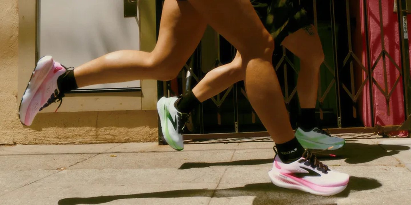 Close-up of two runners wearing Brooks Hyperion Max 2 shoes in pink and green colorways running on a city pavement